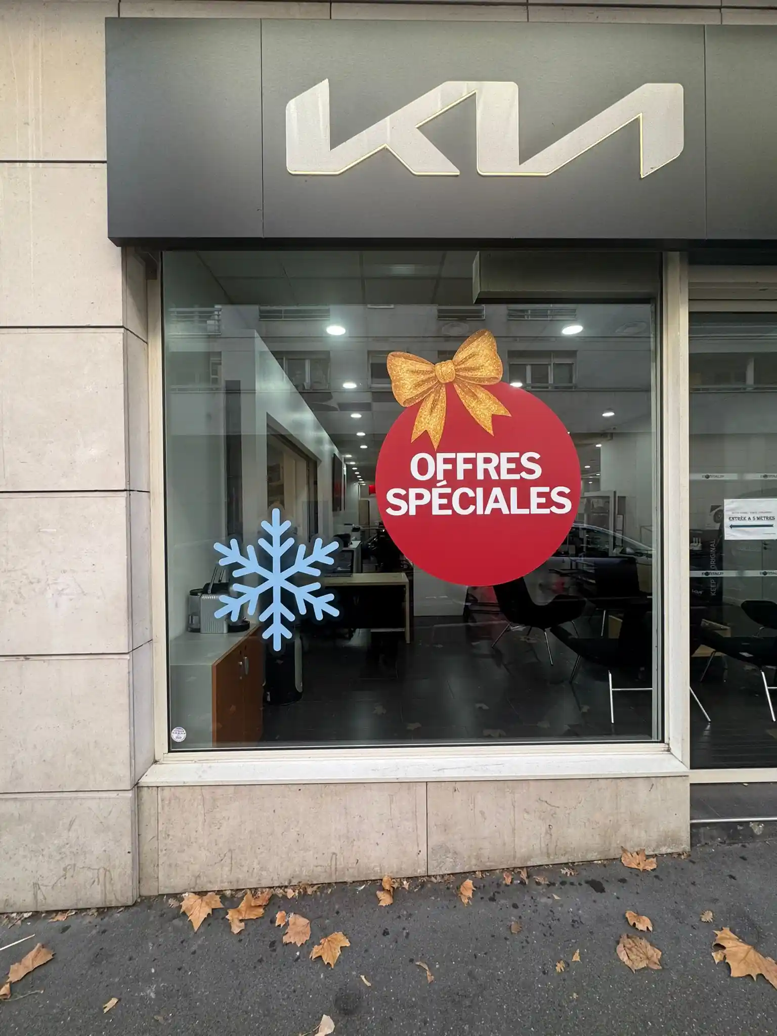 Vitrine de concessionnaire automobile KIA avec une grande vitrophanie de Noël composée d'une boule rouge portant l'inscription blanche 
