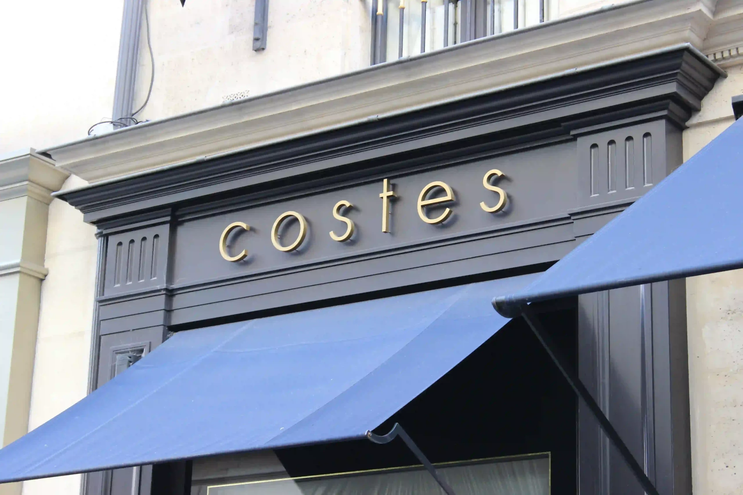Enseigne Costes en lettres découpées dorées sur fond gris anthracite avec un store bannette bleu nuit